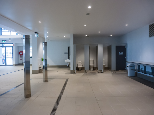 Große öffentliche Toilette mit mehreren Toiletten, tragenden Säulen, einem Mülleimer, einer Tür, einem Schild, einem Feuerlöscher und Deckenleuchten.