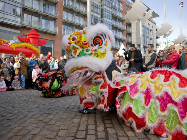 Ein farbenfrohes chinesisches Neujahrsfest in Amsterdam mit einer Löwen-Tanz-Performance vor einer Zuschauermenge, einige halten Kameras, vor dem Hintergrund von Gebäuden, Laternenmasten und einem klaren blauen Himmel.