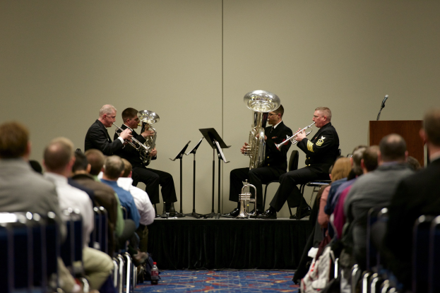 Eine Gruppe von Männern, die auf einer Bühne Blasinstrumente spielen, mit Notenständern, einem Podium mit Mikrofon und einer sitzenden Zuschauermenge.