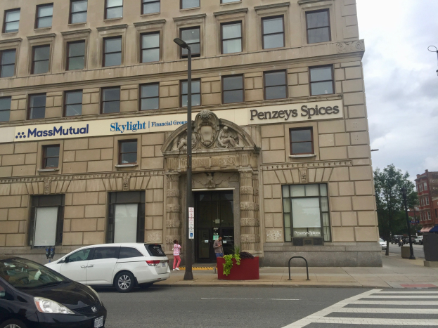 Großes Gebäude mit parkenden Autos, Straßenlaternen, beweglichen Fahrzeugen, Fußgängern, Grünflächen und bewölktem Himmel, identifiziert als Hauptsitz der Massachusetts Mutual Financial Group.