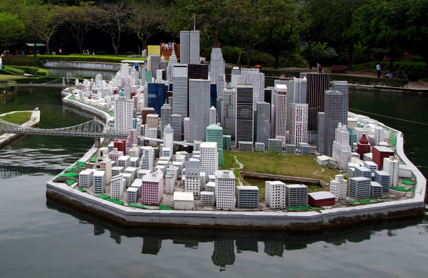 Ein Miniaturmodell einer Stadtlandschaft mit Gebäuden, Grünflächen, einer Brücke und Menschen, das auf einem Gewässer mit einem Himmelhintergrund steht.