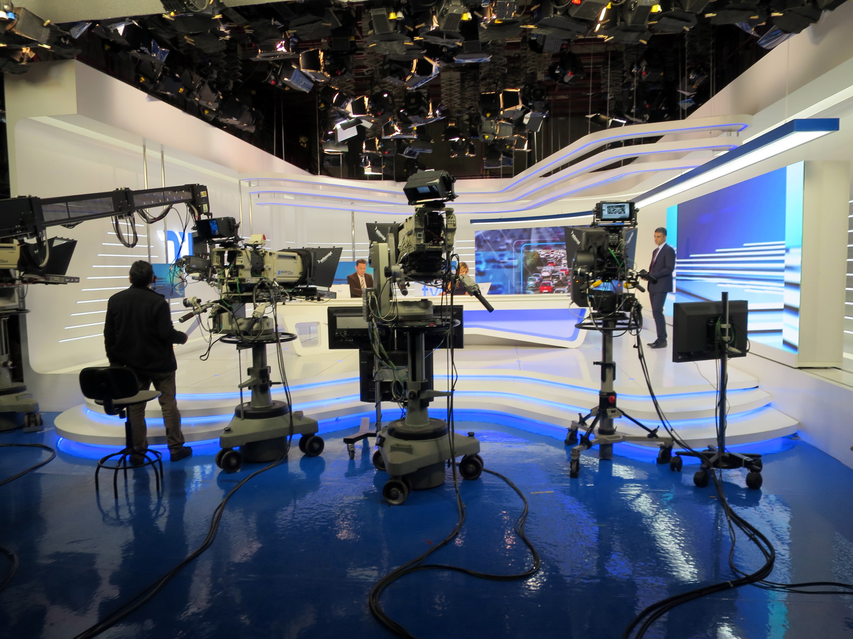 Ein TV-Studio mit mehreren Kameras auf dem Boden, einer sitzenden Person im Vordergrund und einigen stehenden Personen im Hintergrund, beleuchtet von Deckenlampen für eine Live-Übertragung.