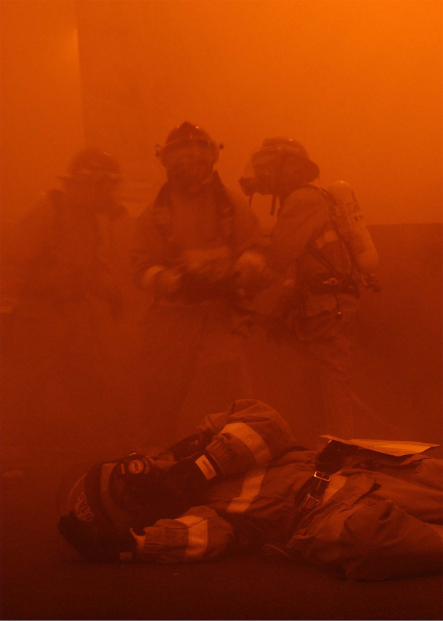 Schwarz-weißes Bild eines Feuerwehrmanns, der vor intensiven Flammen auf dem Boden liegt, umgeben von einer Gruppe von Helmetträgern mit Sauerstoffflaschen in der Nähe.