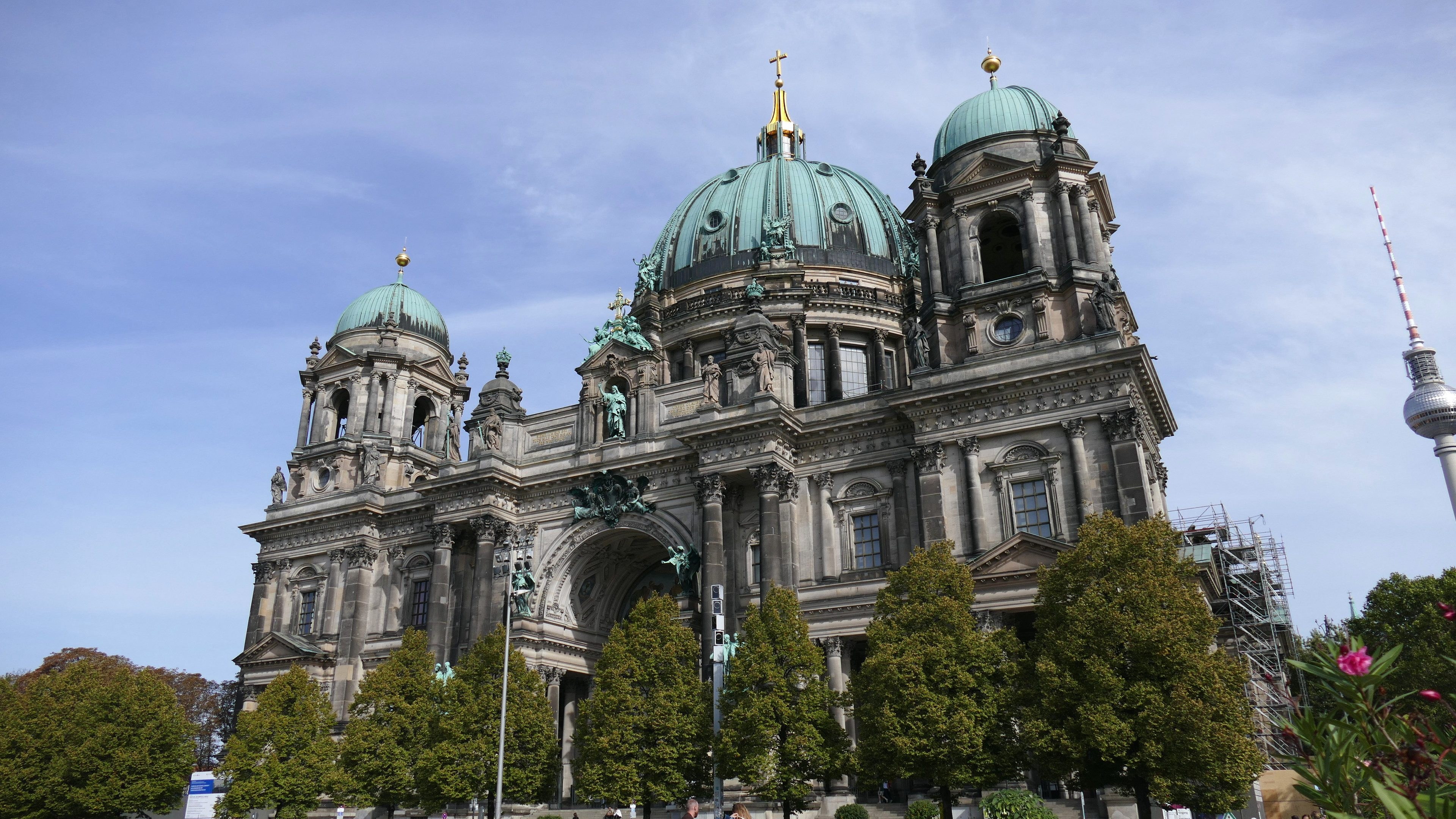 Der Berliner Dom, eine große Kathedrale mit Fenstern, Säulen, Bögen und Statuen, steht prominent in Berlin, Deutschland, mit Menschen vorneweg, Bäumen, Blumenpflanzen und einem Turm in der Nähe unter einem bewölkten Himmel.