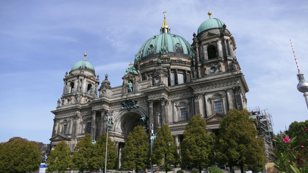 Der Berliner Dom, eine große Kathedrale mit Fenstern, Säulen, Bögen und Statuen, steht prominent in Berlin, Deutschland, mit Menschen vorneweg, Bäumen, Blumenpflanzen und einem Turm in der Nähe unter einem bewölkten Himmel.