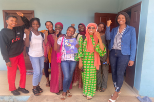 Eine Gruppe von Frauen steht zusammen vor einem Gebäude, lächelt und hält Bücher in den Händen, mit einem Tuch auf der rechten Seite und Türen und einer Wand im Hintergrund, was darauf hindeutet, dass sie Teil einer Schule in Nigeria sind, die über die Bedeutung der Bildung lernt.