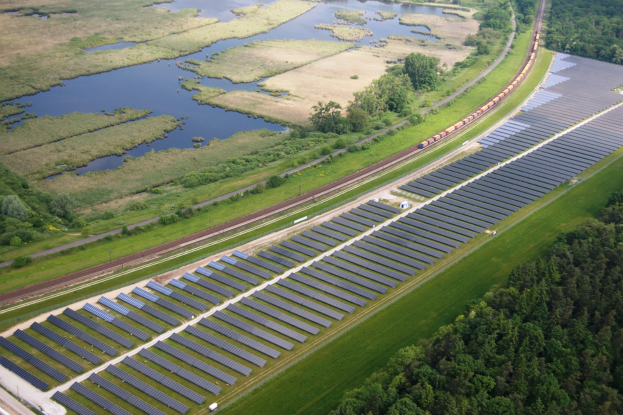 Eine Luftaufnahme eines Solarparks mit zahlreichen Solarpanelen in einem Feld, umgeben von Bäumen, Gras und Wasser, mit einem Zug, der auf einem Bahnsteig in der Nähe fährt.