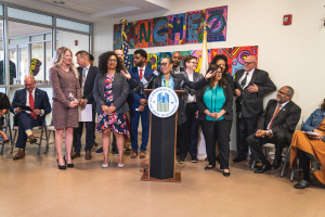 Gruppe von Menschen vor einem Rednerpult mit einem Mikrofon und einer Tafel mit der Aufschrift "Sancho City Councilman Joins City Council", mit sitzenden und stehenden Gästen, Flaggen, Feuerlöscher und Deckenleuchten im Hintergrund.