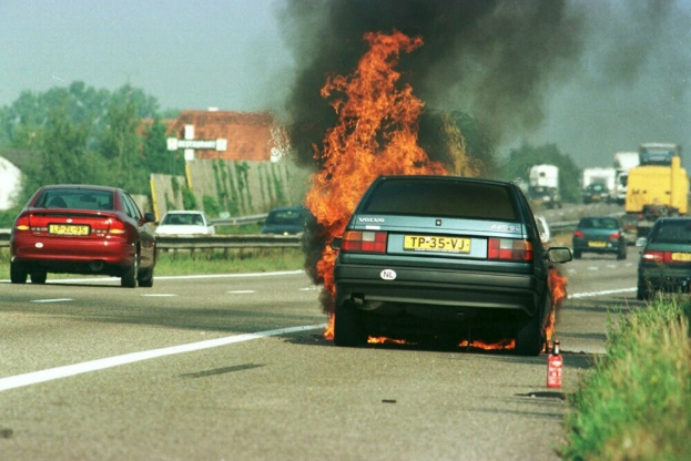 Ein Auto, das in Flammen steht, auf der Seite der Straße, umgeben von anderen Fahrzeugen, mit Bäumen, Gebäuden und einem klaren blauen Himmel im Hintergrund und einem Feuerlöscher auf der rechten Seite des Grases.