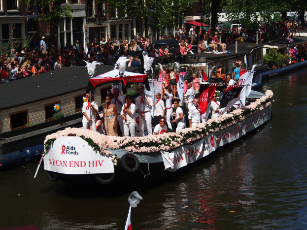 Eine Gruppe von Menschen auf einem Boot im Wasser, umgeben von Fahnen und Blumen, mit Gebäuden, Bäumen und einem Laternenpfahl im Hintergrund während des Amsterdam-Rot-Kreuz-Parades.