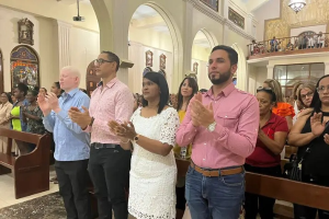 Eine Gruppe von Menschen steht vor einer Kirche und klatscht feierlich, mit Bänken auf beiden Seiten und einem Teppich auf dem Boden, während einige Menschen hinter einem Geländer im Hintergrund stehen.