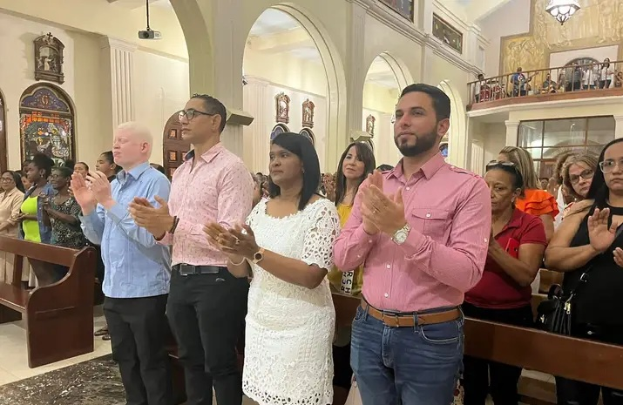 Eine Gruppe von Menschen steht vor einer Kirche und klatscht feierlich, mit Bänken auf beiden Seiten und einem Teppich auf dem Boden, während einige Menschen hinter einem Geländer im Hintergrund stehen.