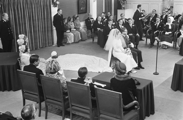 Schwarzes und weißes Foto einer Hochzeitszeremonie mit sitzenden und stehenden Menschen, einem Mann mit Mikrofon und einer Frau mit Blumenstrauß, einem Podium, Blumenschmuck, einem Vorhang, einem gerahmten Foto an der Wand und einer Tür im Hintergrund.