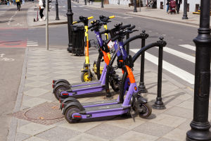 Eine Reihe von Elektro-Scootern, die auf der Straßenseite geparkt sind, mit Menschen, die auf dem Gehweg gehen und Fahrräder fahren, einem Mülleimer im Vordergrund und Gebäuden, Bäumen und Pflanzen im Hintergrund.