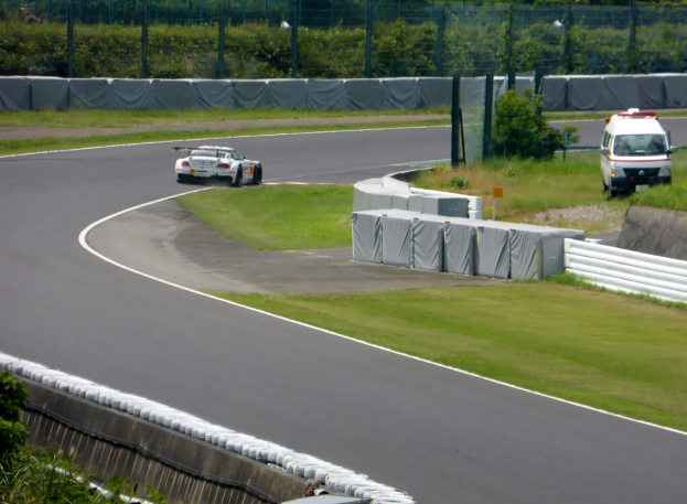 Ein Rennauto fährt auf einer Rennstrecke, im Hintergrund ist ein Polizeiwagen zu sehen, umgeben von Gras, Pflanzen, Bäumen und einem Zaun, mit einem Fahrzeug auf der rechten Seite und Text unten.