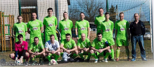 Gruppe von Männer in grünen T-Shirts, die auf einem Fussballfeld stehen oder knien, mit einem Ball und einem Torpfosten umgeben von Bäumen, Gebäuden und Fahrzeugen.