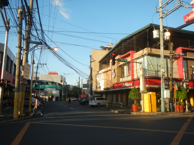 Eine belebte Stadtstraße mit Fußgängern, Fahrzeugen, Versorgungsmasten, Ampeln, Gebäuden mit Schildern, Topfpflanzen und einem bewölkten Himmel.