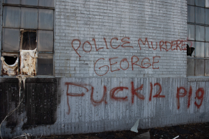 Ein Gebäude mit Graffiti an der Seite, das "Polizei Mord George Fuck 12 P19" liest, mit Fenstern und verschiedenen Gegenständen davor.