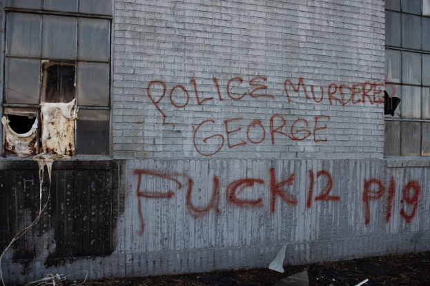 Ein Gebäude mit Graffiti an der Seite, das "Polizei Mord George Fuck 12 P19" liest, mit Fenstern und verschiedenen Gegenständen davor.