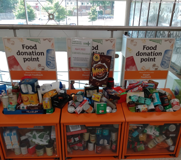 Orangene Schränke mit Lebensmittelspenden gefüllt, darunter Flaschen, Dosen und Kartons, mit Informationsschildern in der Nähe und einem Blick durch Glasfenster auf Fahrzeuge, Bäume, Gebäude und Himmel.