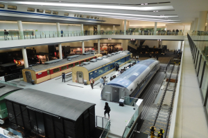 Ein großer Museumsraum mit verschiedenen Bahnen in unterschiedlichen Größen und Farben, mit Menschen, die beobachten, und Geländern, Säulen und Deckenleuchten.