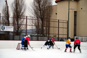 Menschen, die Eishockey auf einem Eisplatz mit Gebäuden, Bäumen, einer Straßenlaterne, einer Namensanzeige und Zäunen im Hintergrund unter einem klaren Himmel spielen.