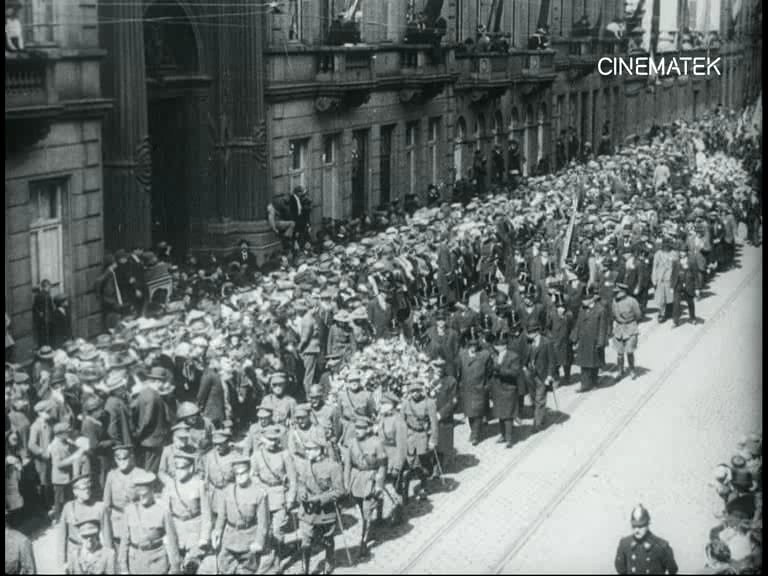 Schwarzes und weißes Foto einer großen Menge, die auf einer Straße marschiert, einige halten Gewehre, vor einem Gebäude mit einem Wasserzeichen in der oberen rechten Ecke.