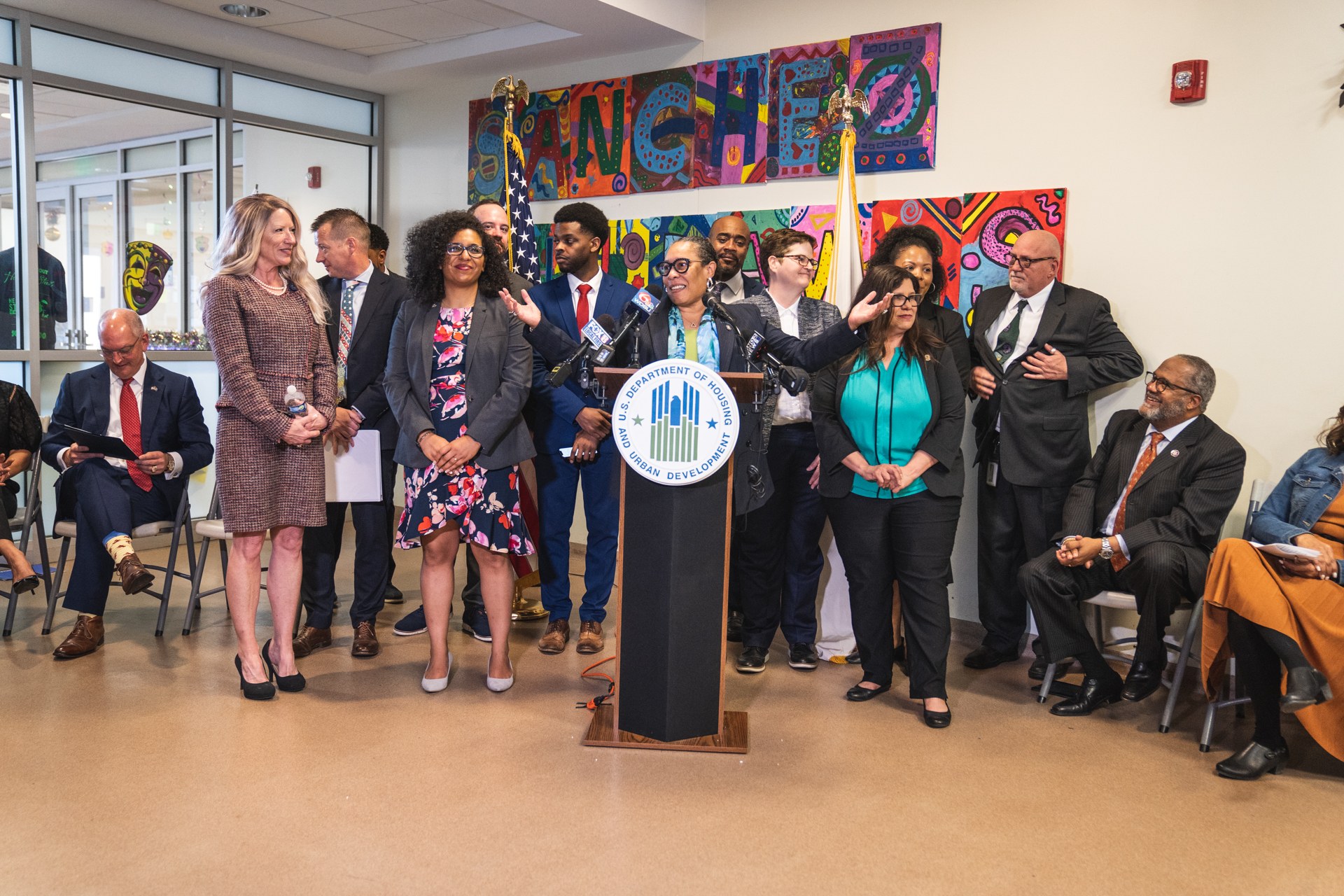 Gruppe von Menschen in einem Raum mit einem Rednerpult, Mikrofon und einer Tafel mit der Aufschrift "Sancho-Stadtrat tritt dem Stadtrat bei", umgeben von Flaggen, Sitzplätzen und einem Feuerlöscher.