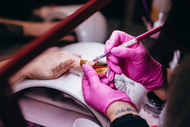 Eine Frau in pinken Handschuhen hält einen Pinsel und eine Nagellackflasche, während sie sich die Nägel in einem Salon machen lässt, mit Maniküre- und Pedikürewerkzeugen auf dem Tisch.