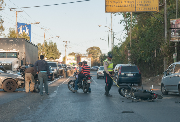 Eine Gruppe von Menschen steht um ein verunglücktes Motorrad am Straßenrand mit mehreren Fahrzeugen, darunter ein Lastwagen, und einem Hintergrund aus Bäumen, Polen, Lampen und Schildern unter dem Himmel.