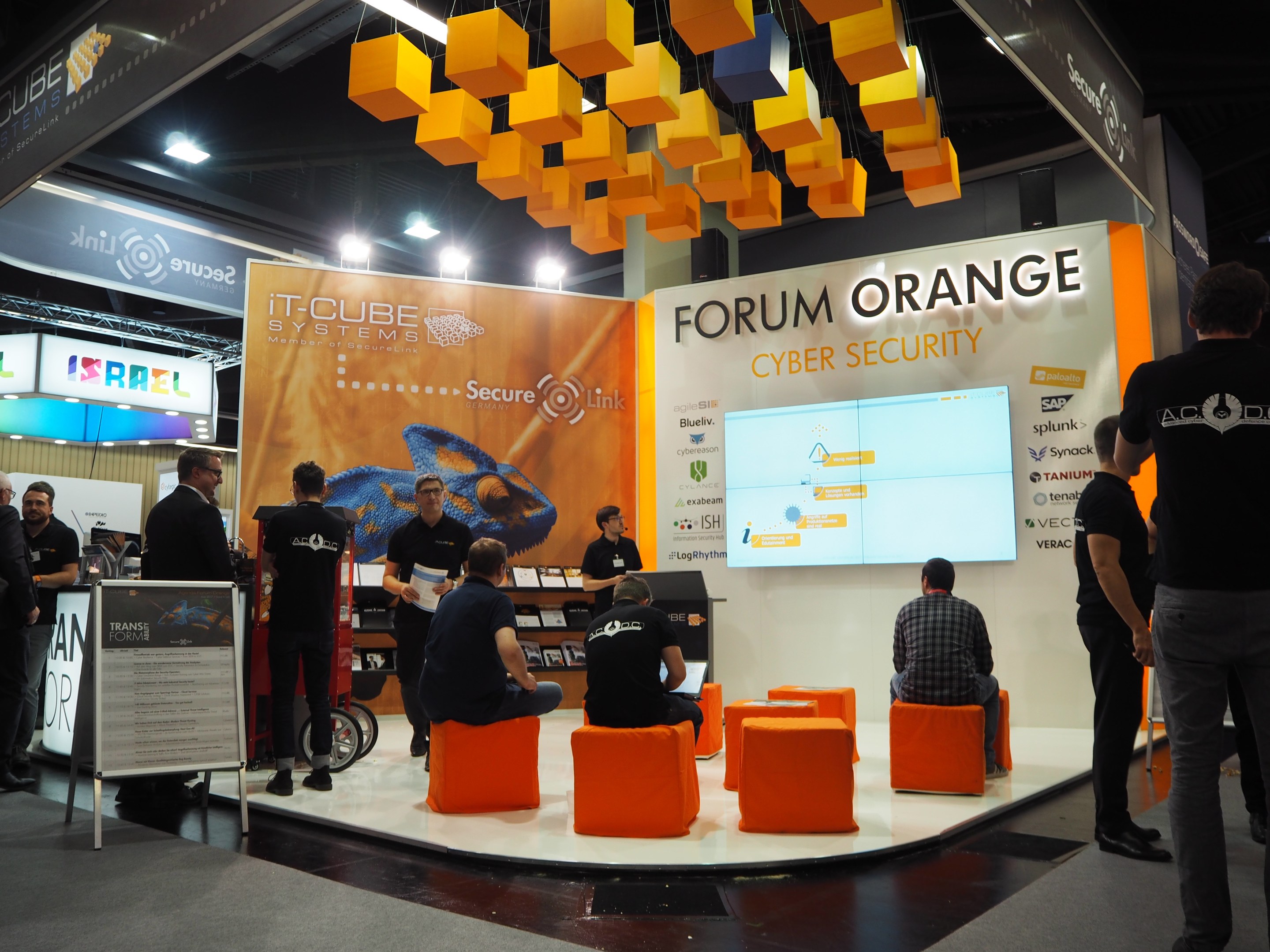 Eine Gruppe von Menschen an einem Messestand für IT-Cube Systems Forum Orange Cyber Security, mit sitzenden und stehenden Teilnehmern, einem Display, Regalen mit Gegenständen, Texttafeln und Deckenleuchten.
