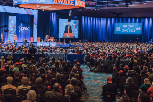 Eine große Menschenmenge sitzt auf Stühlen, die einer Bühne zugewandt sind, mit einigen Personen, die auf dem Podium und auf der linken Seite stehen, blaue Vorhänge und ein Bildschirm im Hintergrund und ein orangefarbenes Schild oben, das die CPAC-Veranstaltung am 25. Oktober anzeigt.