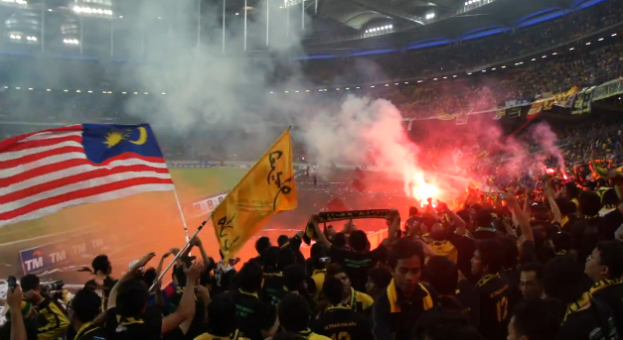Eine große Menge Menschen in einem Stadion mit Fackeln, Fahnen und einer Kamera, mit Rauch und Schildern mit der Aufschrift "Malaysia vs Malaysia Live Streaming" im Hintergrund.