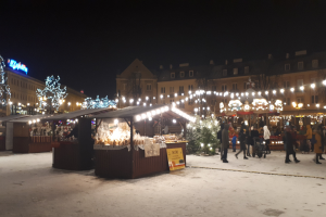 Weihnachtsmarkt in einer verschneiten Straße bei Nacht, mit Menschen, Kinderwagen, beleuchteten Hütten, Pflanzen, Bäumen, Gebäuden und Schildern unter einem bewölkten Himmel.