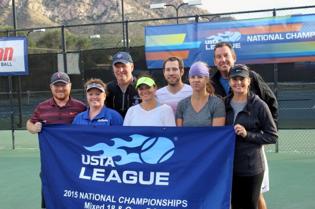 Gruppe von Menschen auf einem Tennisplatz mit einem "2015 USTA National Championships"-Schild, einem Zaun, Schildern, Laternenmasten, Bäumen, Bergen und einem klaren blauen Himmel im Hintergrund.