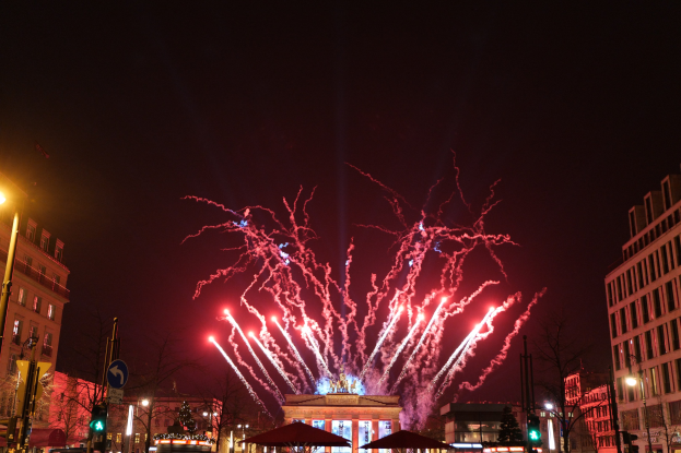 Eine belebte Stadtstraße in Berlin am Silvesterabend, voller Menschen, Fahrzeuge, Bäume, Pfosten, Lichter, Schilder, Verkehrszeichen und Gebäude, beleuchtet von Gebäudelichtern und Feuerwerk am Himmel.