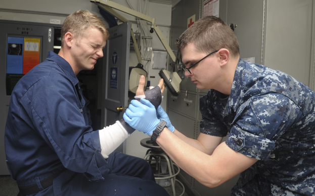 Zwei Männer in militärischer Uniform, einer sitzt mit einem Gips am Arm und der andere steht neben ihm, in einer medizinischen Einrichtung mit medizinischer Ausrüstung im Hintergrund.