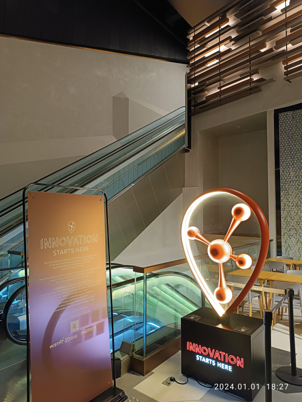 Großes Schild in der Mitte eines Gebäudes mit einer Rolltreppe im Hintergrund, Tische und Stühle rechts und eine Tafel mit "Innovation Startet Hier" links, was auf die HSTP20 Expo hinweist.