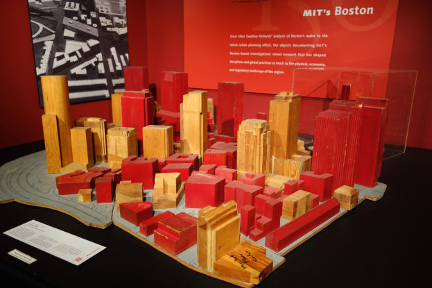 Ein Miniaturmodell einer Stadt aus Holzklötzen auf einem Tisch in einem Museum, begleitet von Papieren, einer Glasbox und einem Rahmenfoto an der Wand.