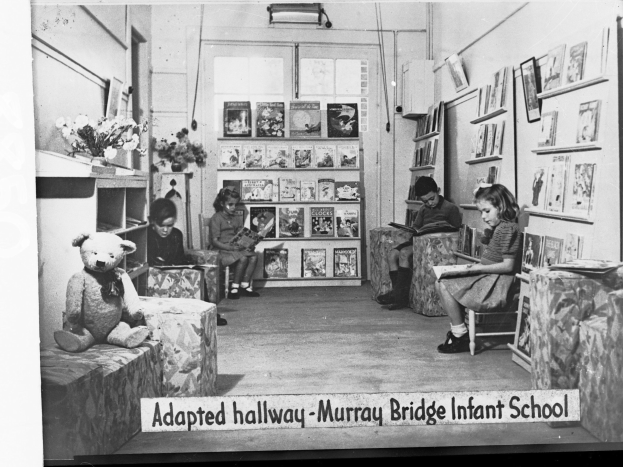 Eine Gruppe von Kindern sitzt auf Stühlen in einem Raum mit Bücherregalen, einem Teddybär links, einer Vase auf dem Tisch und gerahmten Gegenständen an der Wand, mit Text unten: "Angepasster Flur Murray Bridge Kindergarten"