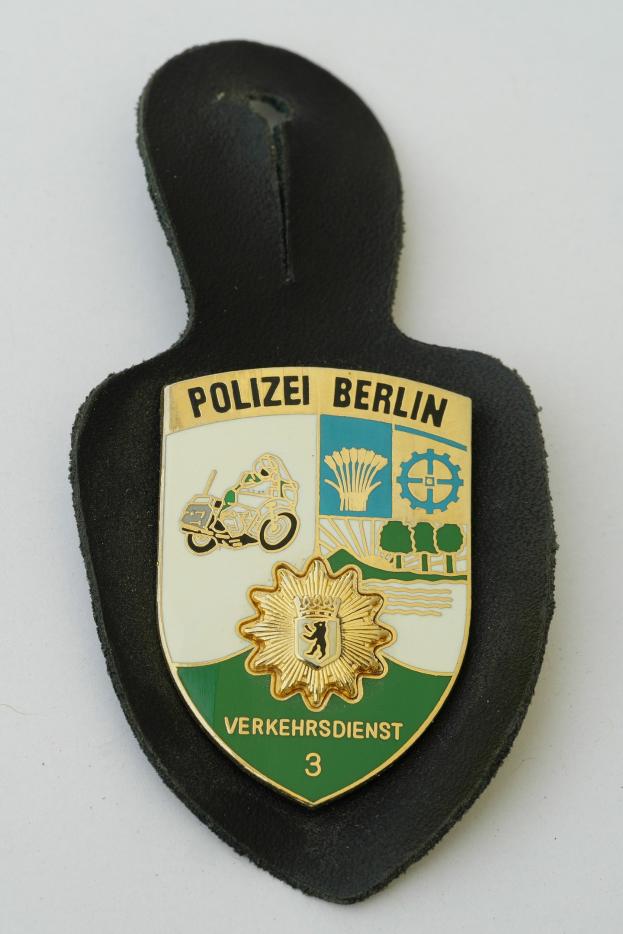 Ein schildförmiges Polizeisiegel mit 'Polizei Berlin' in schwarzer fetter Schrift auf einem blauen Hintergrund mit einem weißen Stern und Rahmen, das auf einer Oberfläche ruht.