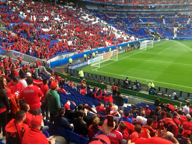 Eine große Menschenmenge in einem Stadion schaut einem Fußballspiel zu, mit Sitzplätzen, Schildern, einem Torpfosten und Deckenleuchten.