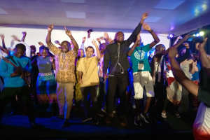 Eine Gruppe von Menschen tanzt auf einer Bühne mit erhobenen Händen, beleuchtet von Scheinwerfern auf der rechten Seite und einem Hintergrundbild.