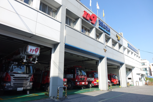 Feuerwache mit mehreren Feuerwehren vor dem Gebäude mit Fenstern, Säulen und Namensschildern, mit Fahnen im Vordergrund und anderen Gebäuden gegen einen klaren blauen Himmel.