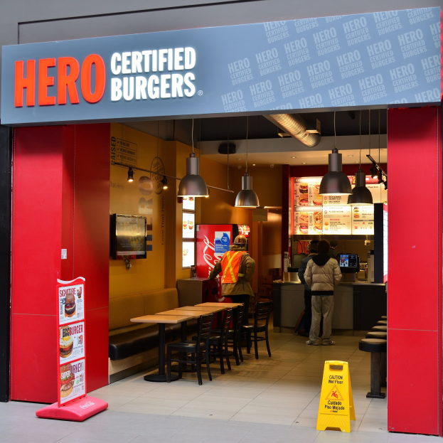 Außenansicht eines Hero Certified Burgers Restaurants in London mit Menschen, Tischen, Stühlen, einer Speisekarte, einem Wandfernseher und Deckenleuchten.