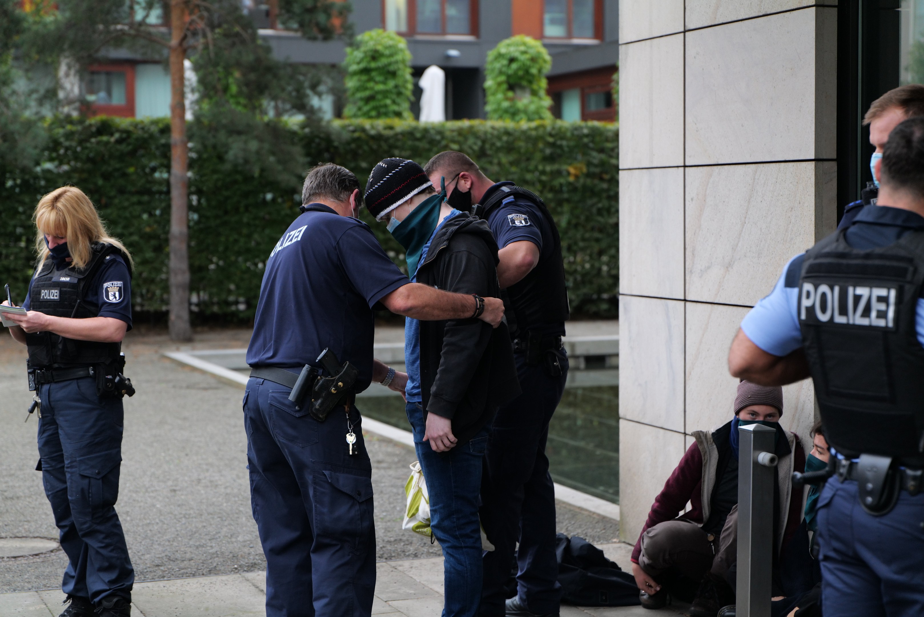 Eine Gruppe von Polizeibeamten steht vor einem Gebäude, während eine Frau ein Buch und einen Stift hält; Menschen sitzen und stehen in der Nähe, mit Bäumen und Gebäuden im Hintergrund.
