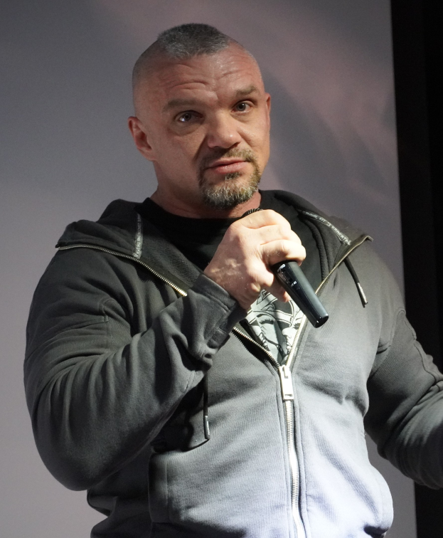 Ein Mann in einer schwarzen Kapuzenjacke und einem schwarzen Anzug mit einem weißen Hemd, zurückgekämmtes Haar und ein selbstbewusstes Ausdruck hält ein Mikrofon, während er vor einer Wand steht.