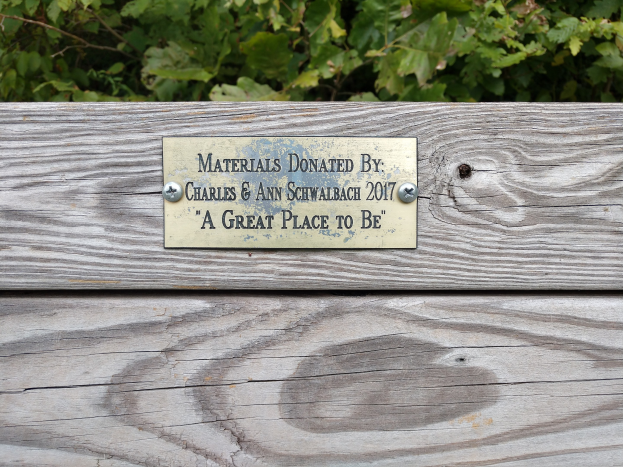 Holzbank mit einer Plakette, auf der "Materialien gespendet von Charles & Ann Schwabach 2017: Ein schöner Ort zu sein" steht, vor üppigen grünen Pflanzen.