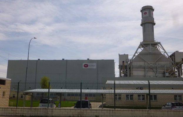 Ein großes Gebäude mit einem zentralen Turm, umgeben von einem Zaun, mit Fahrzeugen auf der benachbarten Straße, Mästen, Lichtern, Drähten, Bäumen und Gras im Vordergrund und einem bewölkten Himmel im Hintergrund, Teil einer neuen Energiespeicherkapazität.
