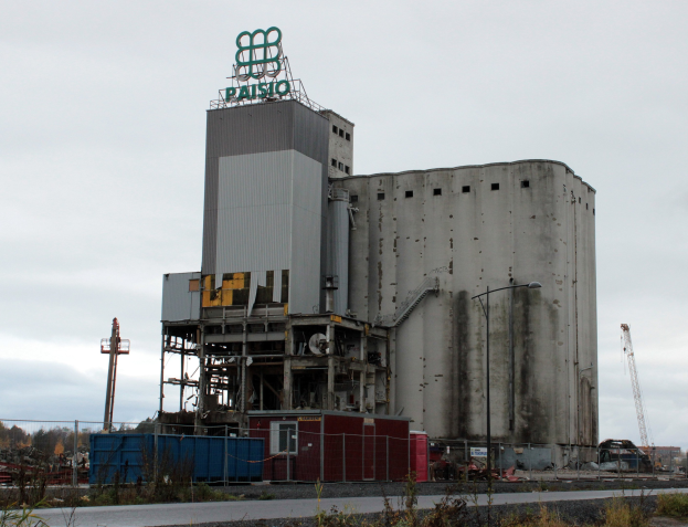 Großes Zementgebäude neben einer Straße, umgeben von Pflanzen, einem Zaun, Pfählen, Lampen und anderen Gegenständen, mit Bäumen und einem klaren blauen Himmel im Hintergrund.
